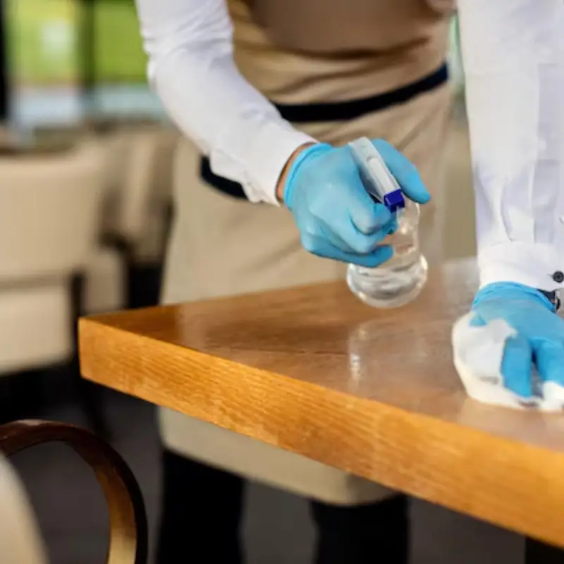 A server in gloves sprays cleaner on a wooden table and wipes it down, ensuring cleanliness in a restaurant setting.