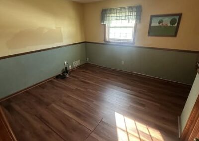 Empty room with hardwood flooring, partially painted walls, a window with green curtains, and a framed painting. Sunlight streams in.