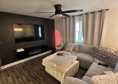 A cozy living room featuring a gray sectional sofa, a large TV mounted on a dark wall, curtains, and wooden flooring.
