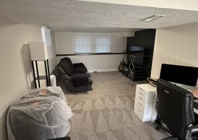 Cozy basement space featuring two recliners, a desk with a computer, and a lamp, all complemented by neat carpeting and bright windows.