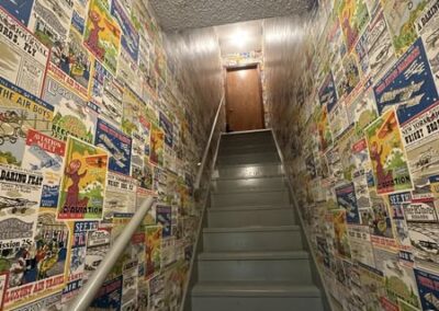 A staircase with a painted gray floor, surrounded by vibrant vintage posters on the walls, leading to a lighted doorway at the top.